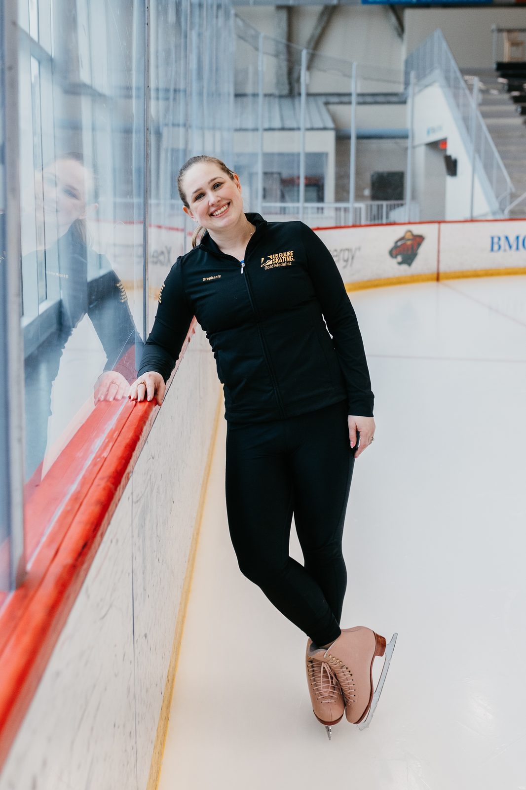 Stephanie Culver on ice in coaching jacket, leaning on the boards at a Twin Cities rink